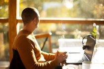 Uomo d'affari premuroso con caffè e laptop guardando fuori dalla finestra soleggiata — Foto stock