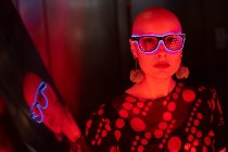Portrait cool woman with shaved head and neon glasses in red light — Stock Photo