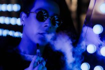 Portrait cool young man in sunglasses vaping in dark neon. nightclub — Stock Photo