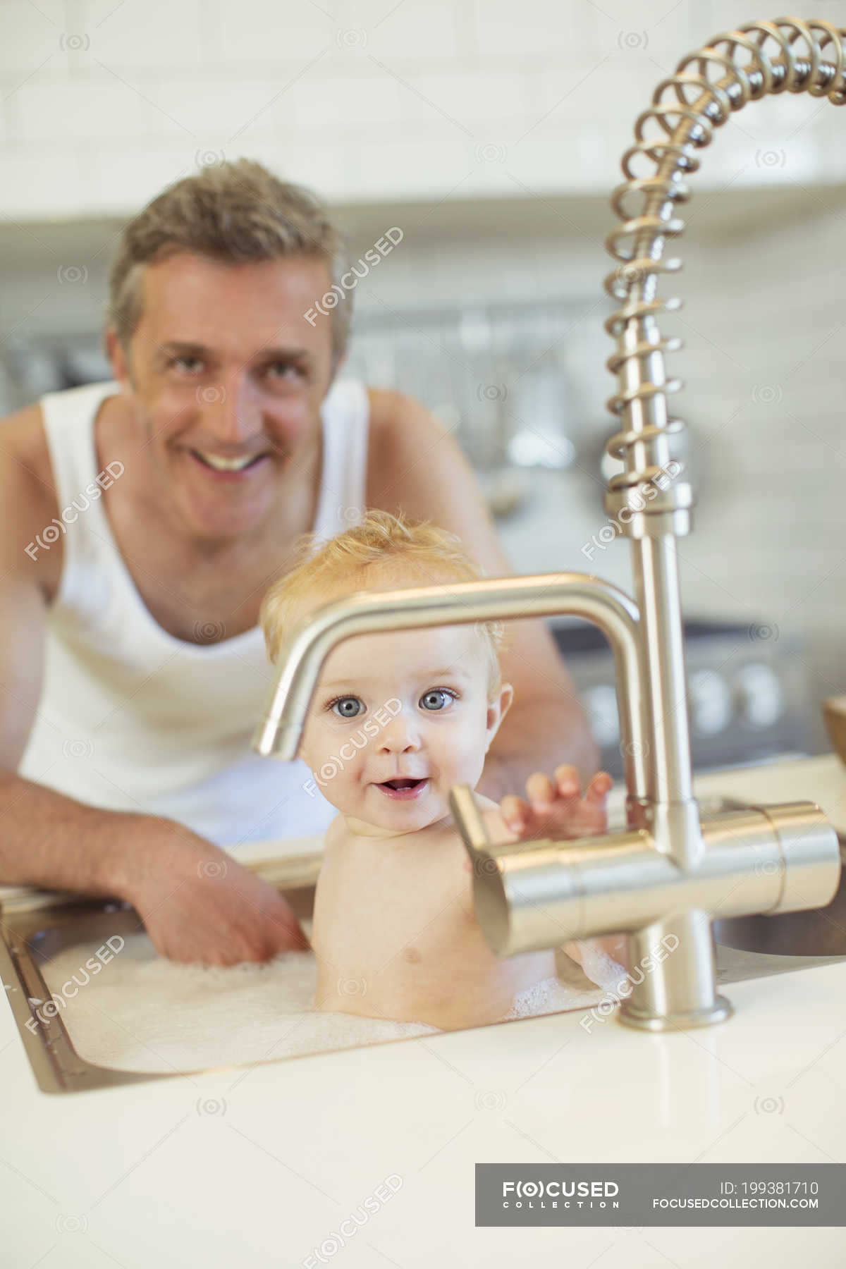Father Bathing Baby In Kitchen Sink Looking At Camera Two