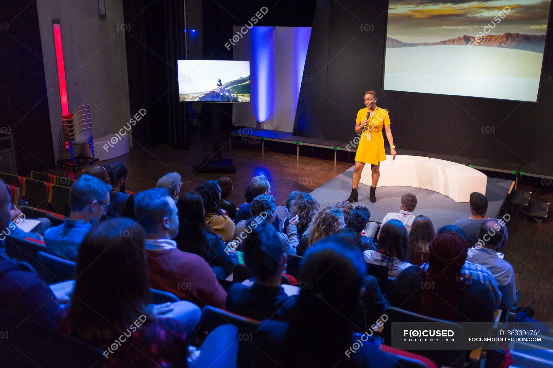 Female speaker on stage talking to audience — auditorium, vanguardians
