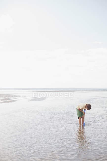 Boy with shovel playing in ocean surf on summer beach — Stock Photo