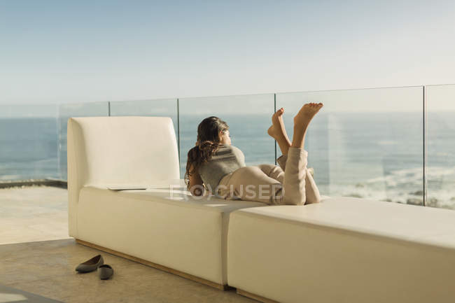 Женщина на роскошном балконе отдыхает лежа на скамейке, глядя на солнечный вид на океан — стоковое фото