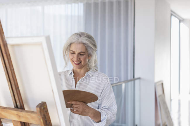 Sorrindo mulher madura com pintura de paleta em tela no cavalete — Fotografia de Stock