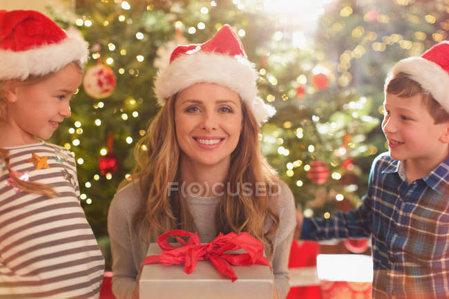 Retrato sonriente madre en Santa sombrero recibiendo regalo de Navidad de la hija y el hijo - foto de stock