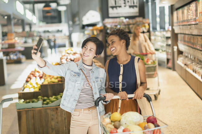 Sonriendo joven pareja de lesbianas tomando selfie en el mercado de comestibles - foto de stock