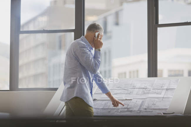 Architect talking on cell phone reviewing blueprints in office — Stock Photo