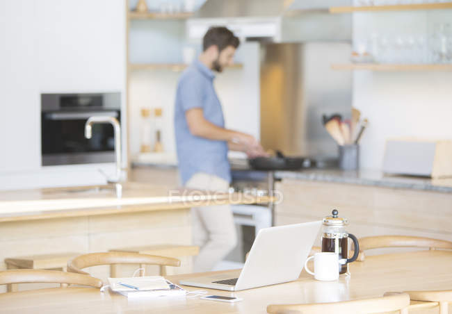 Homme cuisine à la cuisinière derrière ordinateur portable, café et téléphone cellulaire dans la cuisine — Photo de stock