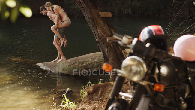 Joven pareja piggybacking en lakeside detrás de la motocicleta - foto de stock