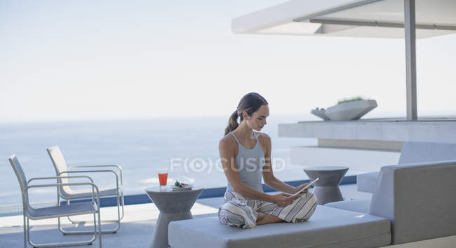 Femme en pyjama utilisant une tablette numérique sur la maison moderne et luxueuse vitrine patio extérieur avec vue sur l'océan — Photo de stock