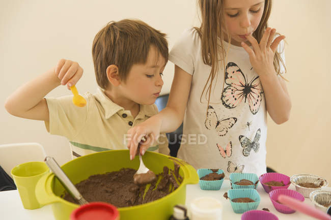 Chico y chica hermano y hermana haciendo cupcakes de chocolate, lamiendo dedos - foto de stock