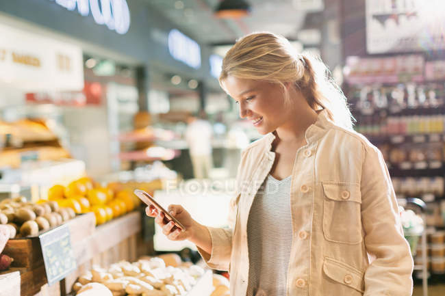 Mujer joven usando el teléfono celular en el mercado de comestibles - foto de stock