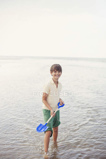 Portrait smiling boy with shovel in ocean surf on summer beach — Stock Photo