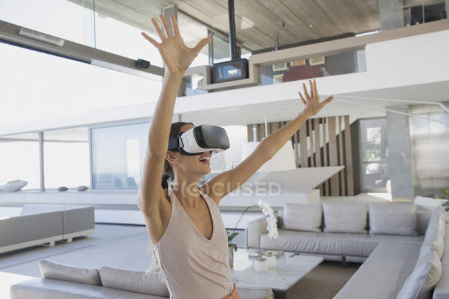 Femme énergique utilisant des lunettes de simulateur de réalité virtuelle avec les bras levés dans le salon moderne et luxueux de vitrine de la maison — Photo de stock