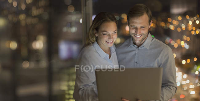 Улыбающийся бизнесмен и деловая женщина, работающая допоздна за ноутбуком в офисе ночью — стоковое фото