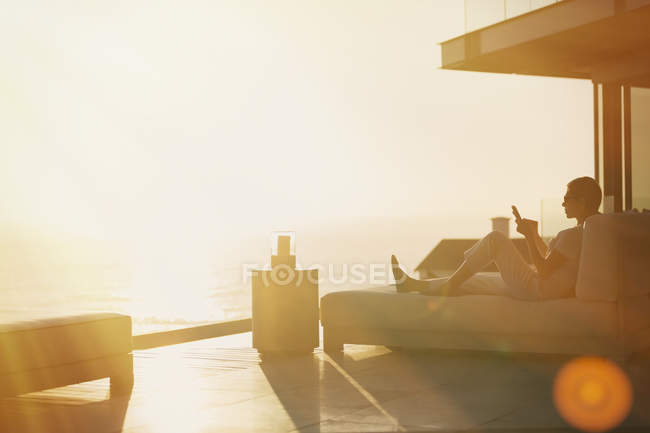 Silueta mujer usando el teléfono celular en el chaise lounge en el balcón de lujo con vista al mar puesta de sol - foto de stock