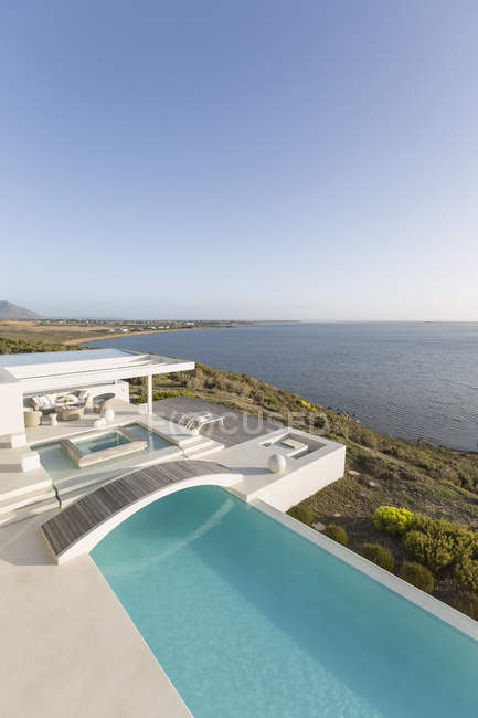 Sunny, tranquille maison de luxe moderne vitrine piscine à débordement avec passerelle et vue sur l'océan sous le ciel bleu — Photo de stock