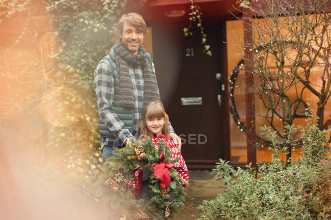 Retrato sonriente padre e hija sosteniendo corona de Navidad fuera de casa - foto de stock