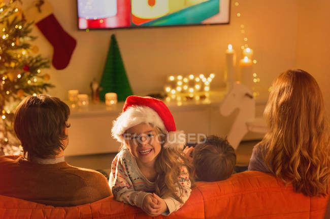 Портрет улыбающейся девушки в шляпе Санты, смотрящей телевизор с родителями в рождественской гостиной — стоковое фото