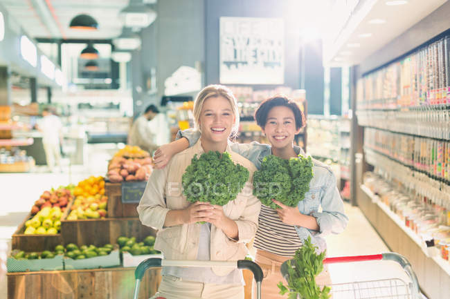 Retrato sonriente jóvenes amigas sosteniendo col rizada en el mercado de comestibles - foto de stock