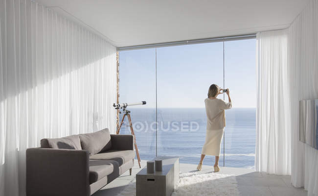 Femme avec appareil photo téléphone photographiant ensoleillé vue sur l'océan de moderne, maison de luxe vitrine salon intérieur — Photo de stock