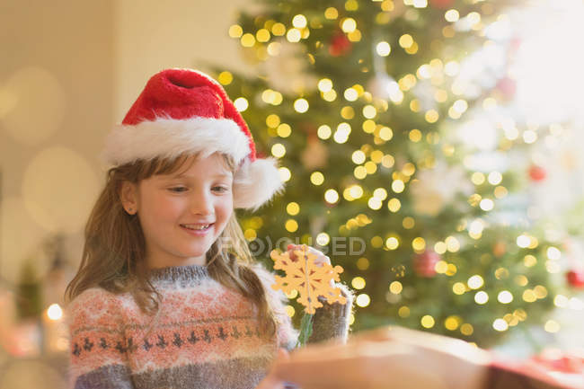 Girl in Santa hat holding snowflake ornament in front of Christmas tree — Stock Photo
