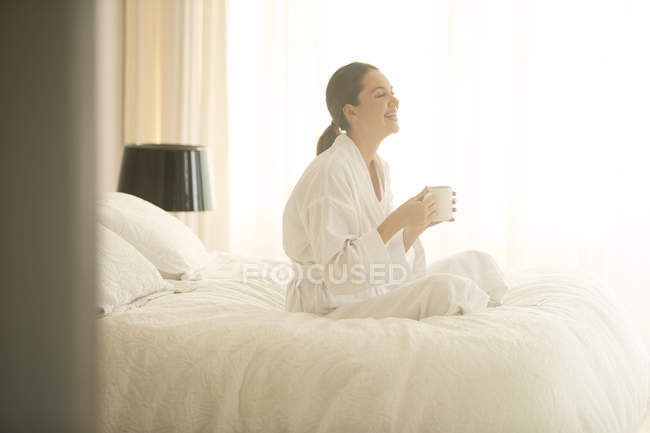 Smiling woman in bathrobe drinking coffee cross-legged on bed — Stock Photo