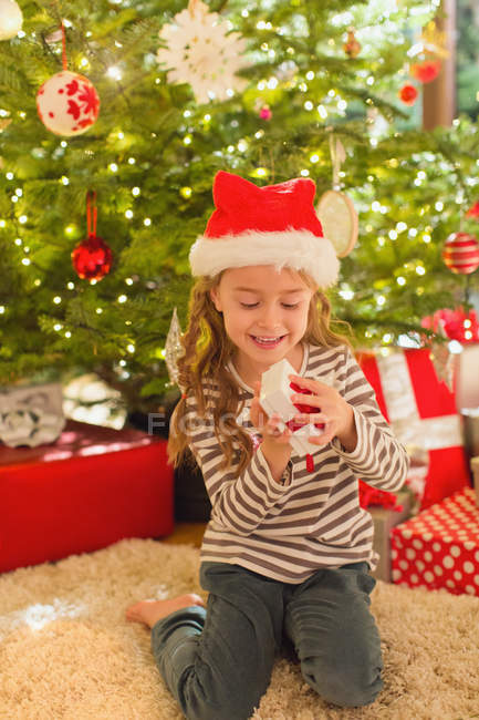 Muchacha sonriente en el regalo de apertura del sombrero de Santa delante del árbol de Navidad - foto de stock