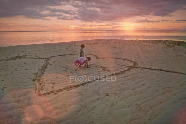 Boy and girl brother and sister drawing heart-shape in sand on tranquil sunset beach — Stock Photo