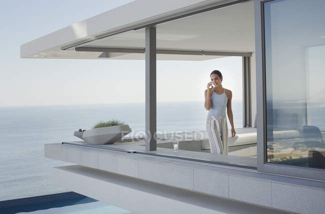 Donna che parla al telefono cellulare sul moderno, lusso casa vetrina patio esterno con vista sull'oceano — Foto stock
