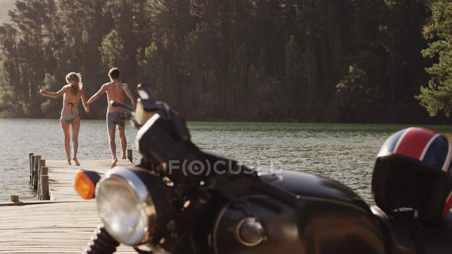 Young couple holding hands and running on lakeside dock behind motorcycle — Stock Photo