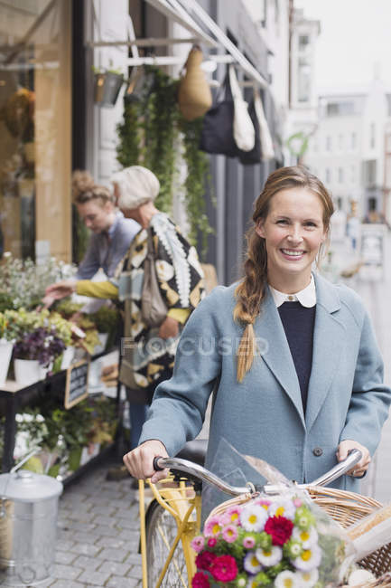 Портрет усміхненої жінки, що йде на велосипеді з квітами в кошику за межами магазину — стокове фото