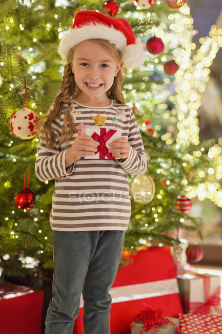 Портрет усміхненої дівчини в капелюсі Санта тримає подарунок перед ялинкою — стокове фото