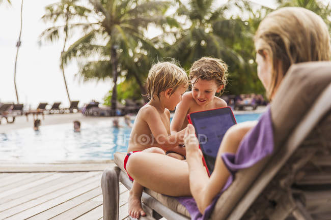 Madre e hijos relajándose con la tableta digital junto a la piscina - foto de stock