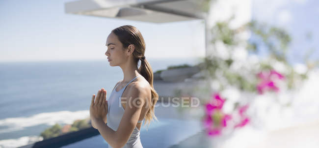 Serene brunette woman practicing yoga, meditating with hands at heart center on sunny patio with ocean view — Stock Photo