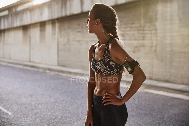 Tired fit female runner in sports bra with mp3 player armband and headphones on urban street — Stock Photo