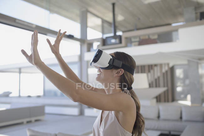 Femme souriante utilisant des lunettes de simulateur de réalité virtuelle avec les bras levés dans le salon moderne et luxueux de vitrine de la maison — Photo de stock