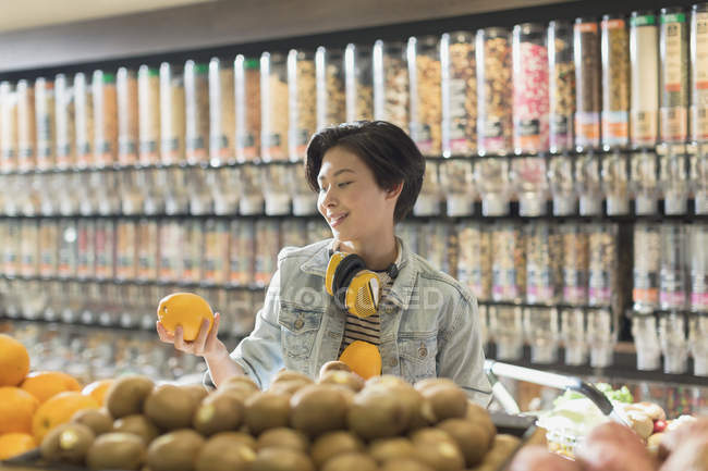 Mujer joven con auriculares compras de comestibles, la celebración de naranja en el mercado - foto de stock