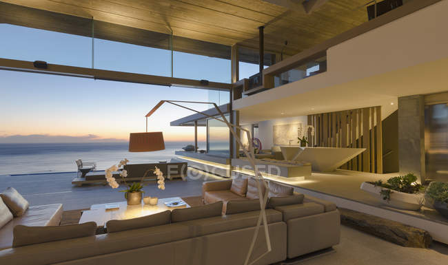 Illuminated modern, luxury home showcase interior living room with ocean view at dusk — Stock Photo