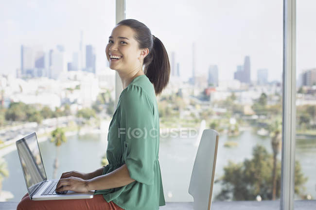 Portrait of confident casual businesswoman at laptop near window overlooking city — Stock Photo