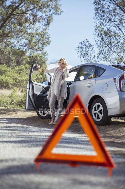 Femme parlant sur un téléphone portable au bord de la route derrière un triangle d'avertissement — Photo de stock