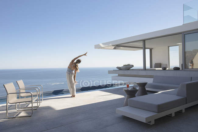 Woman practicing yoga side stretch on modern, luxury home showcase exterior patio with sunny ocean view — Stock Photo