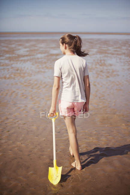 Teenage girl dragging shovel in wet sand on sunny summer beach — Stock Photo