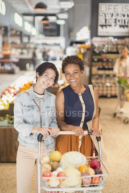 Retrato sonriente joven pareja lesbiana con carrito de la compra compras de comestibles en el mercado - foto de stock