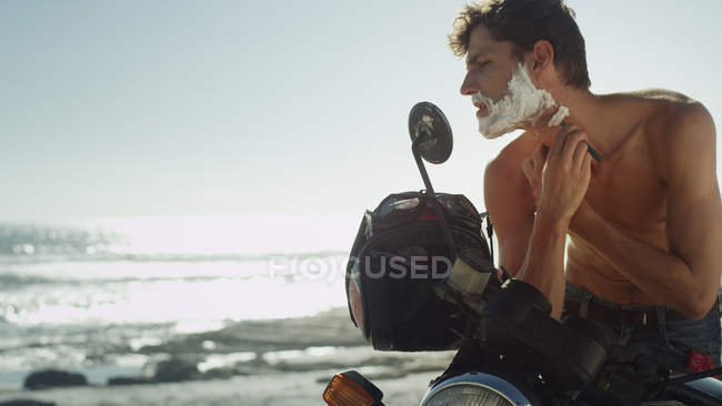 Junger Mann auf Motorrad rasiert Bart in der Nähe des Ozeans — Stockfoto