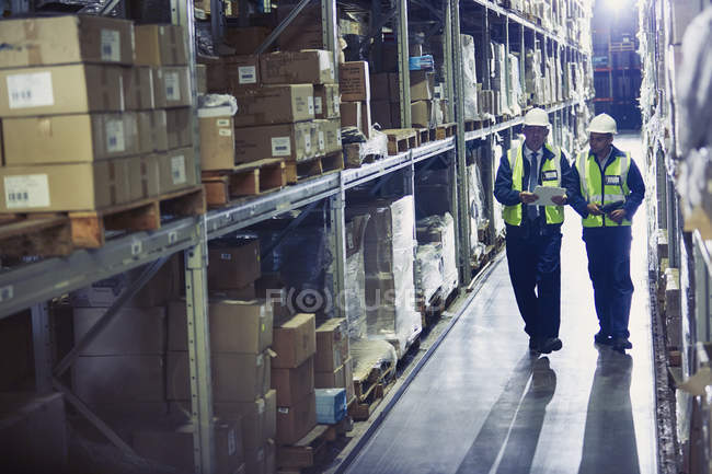 Gerente e trabalhador com prancheta andando ao longo de mercadorias em prateleiras no armazém de distribuição — Fotografia de Stock