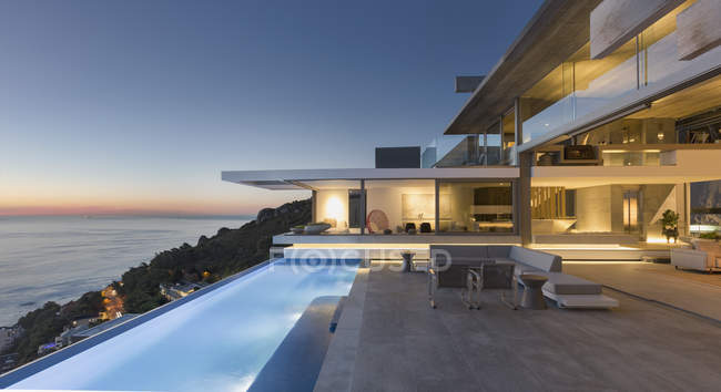 Iluminado moderno, casa de luxo vitrine pátio exterior com piscina de colo e vista para o mar no crepúsculo — Fotografia de Stock