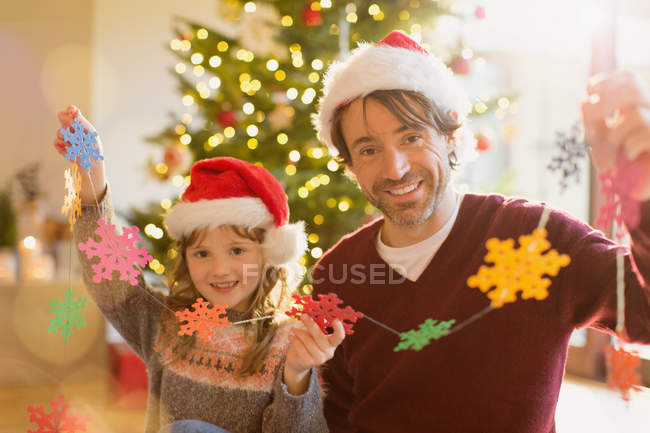 Retrato sonriente padre e hija en sombreros de Papá Noel sosteniendo una cadena de copos de nieve de papel - foto de stock