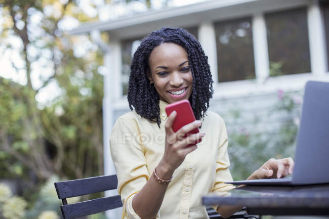 Улыбающиеся женщина, используя мобильный телефон и ноутбук на патио — стоковое фото