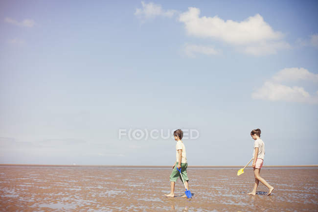 Брат і сестра з лопатами мокрого піску на пляжі сонячних літніх нижче синього неба — стокове фото